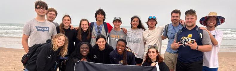 A group of individuals pose on the beach holding a flag with the ETBU logo on it